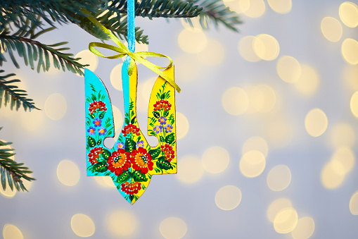 Ukrainian Christmas ornament tryzub. Wooden decoration with Petrykivka painting hanging from a tree with golden lighting with bokeh in the background. Hand painted decor for Christmas tree