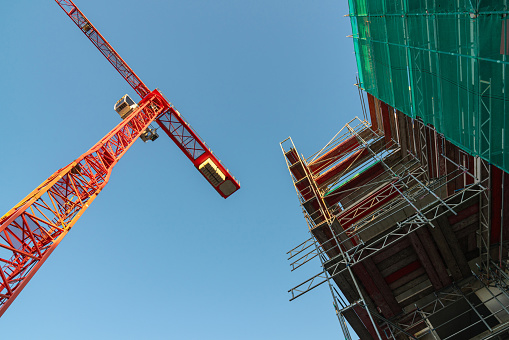 Bottom view of a tower crane and a house under construction surrounded by scaffolding against a blue sky.