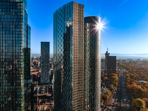 An aerial photograph of Deansgateskyscrapers in downtown Manchester, England. The photograph was produced on a very cold but beautifully bright day, with clear blue skies during autumn. Autumnal trees can be seen at the bottom of the photograph with the sun gleaming off the top of a skyscraper