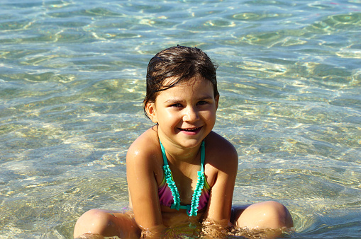 A cheerful five year old girl is sitting on the beach in the shallow sea and smiling in the camera