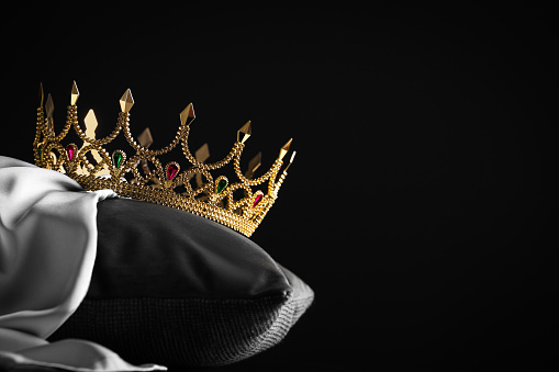 Beautiful gold crown with gems on pillow against black background. Space for text
