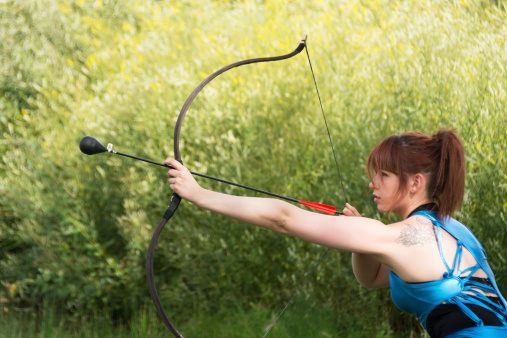 Horizontal outdoor shot of serious young woman aiming practice arrow in profile.