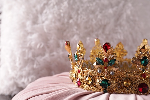 Beautiful golden crown with gems on pink cloth, closeup. Space for text