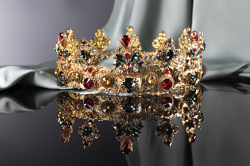 Beautiful golden crown with gems near light cloth on dark mirror surface