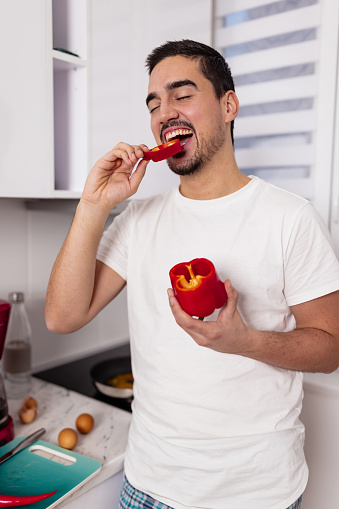 The photo captures a young man in his kitchen holding a fresh paprika, enjoying its crisp taste as a wholesome snack
