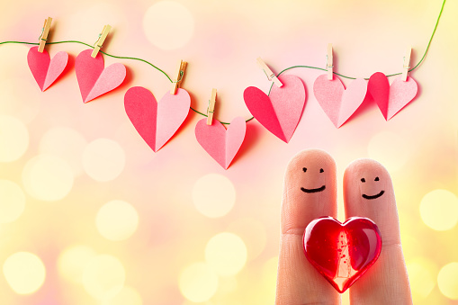 Funny finger couple with a red heart and red paper cut hearts hanging on a clothesline with wooden clothespins. Beautiful defocused lights bokeh background. Space for copy.