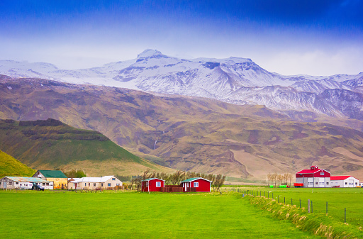 Towering snow capped mountains with cascading waterfalls rise behind Icelandic farmlands on Iceland's south coast.