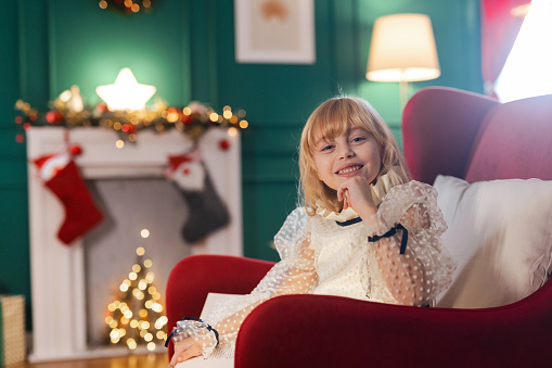 Portrait of a beautiful little girl sitting on the armchair in a living room with Christmas decorations and looking at the camera