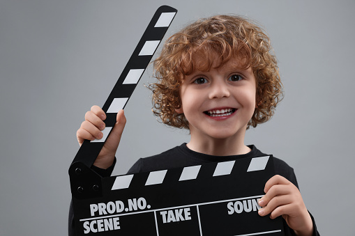 Smiling cute boy with clapperboard on grey background. Little actor