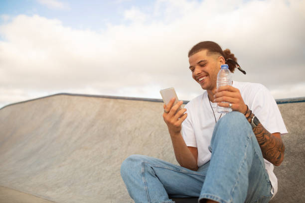 istock Skateboarder using cellphone during break at skatepark and holding bottle with water 1793734015