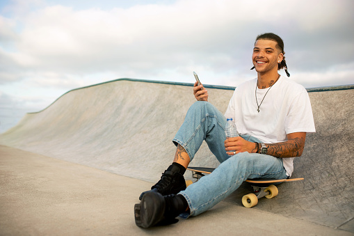 Happy stylish european man using mobile phone while sitting on skateboard on sports ramp, guy smiling at camera, free space. Young skater man enjoying active sports outside