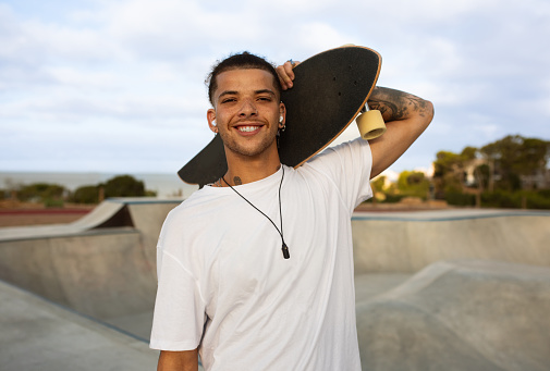 Portrait of happy hipster tattooed european man in white t-shirt posing with skateboard, walking in skate park and smiling at camera, free space