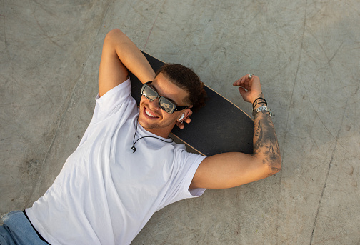 Stylish tattooed man relaxing on skateboard and listening music in earpads, having rest after skateboarding exercises, top view, free space. Guy relaxing in skate park