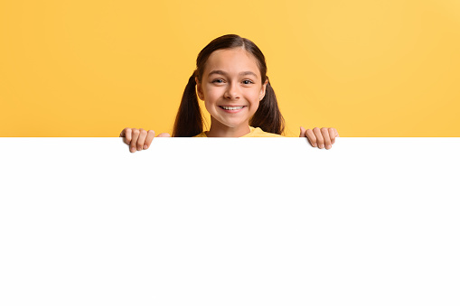Portrait of smiling teen girl hiding behind blank white placard, happy kid demonstrating copy space for your text or design, showing horizontal empty board, standing over yellow background, mockup
