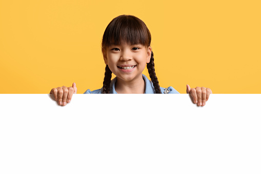 Cheerful cute pretty little asian girl preteen kid posing behind horizontal blank white advertising board, smiling at camera, showing nice advertisement, copy space for text, yellow background