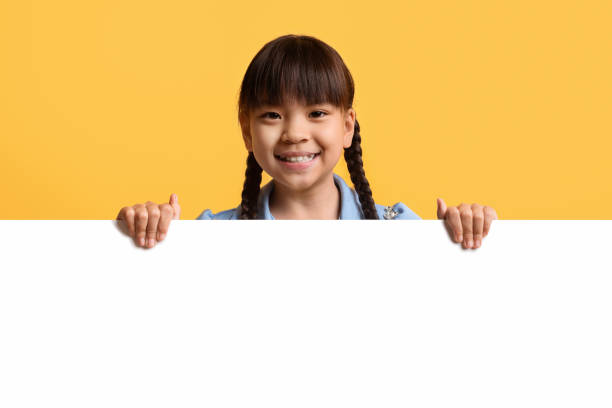 Cute little asian girl kid posing behind blank advertising board Cheerful cute pretty little asian girl preteen kid posing behind horizontal blank white advertising board, smiling at camera, showing nice advertisement, copy space for text, yellow background kids using smart board stock pictures, royalty-free photos & images