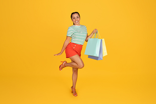 Beautiful cheerful young brunette lady in stylish outfit holding colorful shopping bags, posing isolated on yellow studio background. Happy millennial woman enjoying shopping, showing purchases