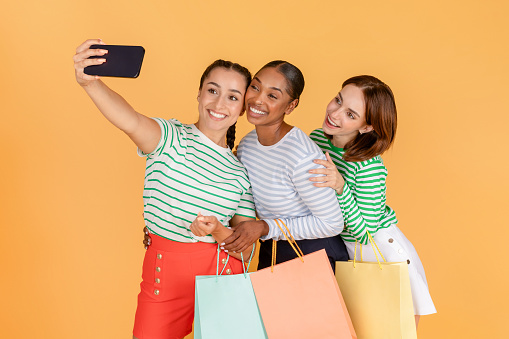 Cute pretty young multiracial ladies shopaholics taking selfie together on cell phone while enjoying shopping, isolated on orange background. Leisure, retails, happy lifestyle concept