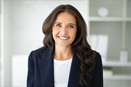 Portrait of cheerful attractive mature woman 50s wearing formal suit leader posing at office, smiling at camera. Closeup photo of happy business lady at workplace. Story of success, women in business