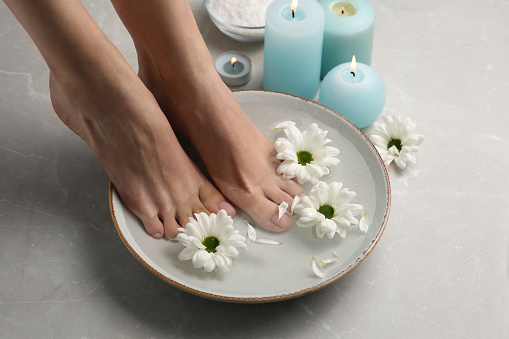 Woman soaking her feet in bowl with water and flowers on grey marble floor, closeup. Pedicure procedure