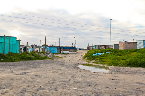 Here is a view from the gravel road of shacks in South Africa. Make-shift buildings are made of corrugated metal, wire fences and wood scraps. Khayelitsha is a suburb of Cape Town. The ground is covered with gravel, dirt, debris, and a puddle of water and mud. See tall tree branches and trucks used for connecting electricity.