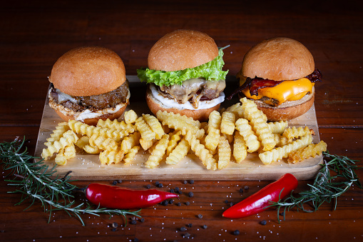 Three burger sliders placed on a wooden board. Very tasty burgers with melted cheese, cheddar, bacon, lettuce, tomato, onion and sauce. Craft beef burgers and french fries, hand made.