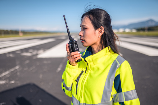 Asian female airfield operations officer monitoring an airfield. Using a walkie talkie to communicate with an airplane and air traffic control.