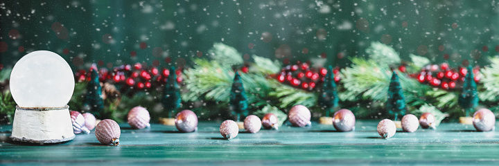 Banner of a magical empty snowglobe over a green rustic table background with falling snow. Christmas decorations in the background. Blurred foreground and background. Copy space available.