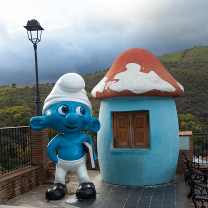 Júzcar, Spain - 3rd November 2023: A Smurf figure outside its mushroom home in Júzcar, Serranía de Ronda. Formerly one of Andalucía's White Villages, it was painted blue by Sony to promote their movie The Smurfs in 2011. A subsequent village ballot voted to retain the blue colouring because of increased tourism, and the village now features various Smurf-related activities.