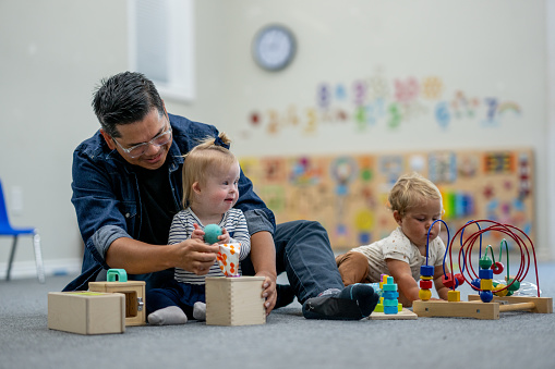 A male caregiver sits on the floor with some toddlers in a daycare classroom as they learn through play.   One little girl with Down Syndrome is seated in closely as she discovers various toys with the caregiver.