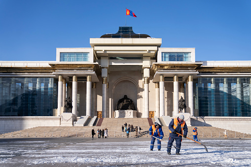Ulaanbaatar, Mongolia - November 8, 2023: People walk around a snow-covered Sukhbaatar Square, in front of an equestrian statue of Damdin Sükhbaatar and the Government Palace.