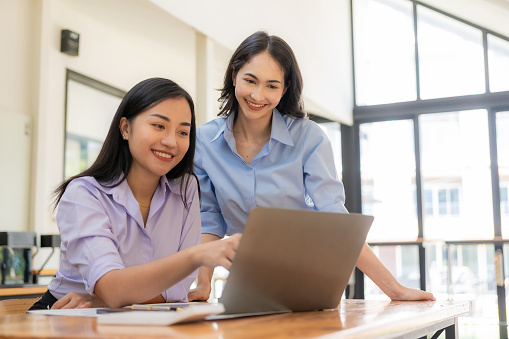 Two Asian women working with laptops and financial documents are discussing marketing plans. The new company's sales strategy is in line with current events.