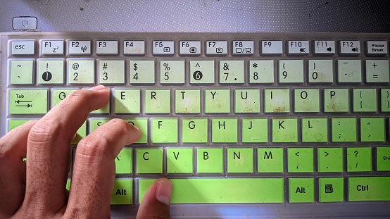 Close up image of a man's hands typing on a laptop keyboard and surfing the internet at an office desk, with a variety of green and white keyboard colors