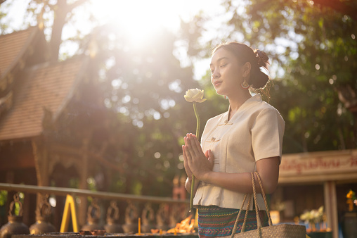 Side view image of a beautiful Asian woman in a traditional Thai-Lanna dress with a lotus flower in her hands is making a wish in a temple. praying, paying respect to the Buddha, Thai culture