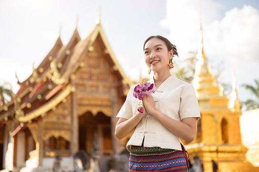 A beautiful Thai woman in a traditional Thai-Lanna dress with a garland is putting her hands together in a prayer position while standing in front of a beautiful chapel in a temple. make a wish
