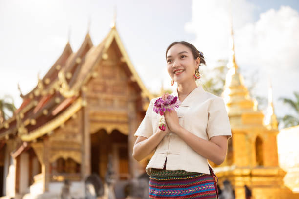 A beautiful Thai woman in a Thai-Lanna dress is putting her hands together in a prayer position. A beautiful Thai woman in a traditional Thai-Lanna dress with a garland is putting her hands together in a prayer position while standing in front of a beautiful chapel in a temple. make a wish traditional thai costume stock pictures, royalty-free photos & images