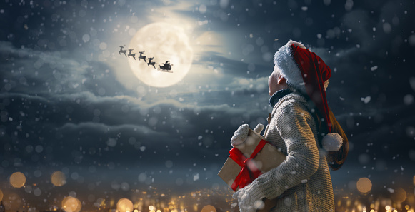 Merry Christmas. Cute little child with xmas present. Santa Claus flying in his sleigh against moon sky. Happy kid enjoy the holiday. Portrait of girl with gifts on dark background.