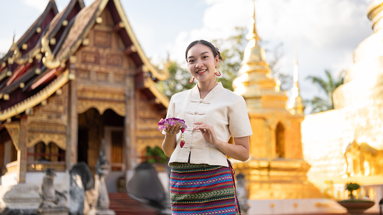 A beautiful and charming Thai woman in a traditional Thai-Lanna dress with a garland in her hand is standing in front of a beautiful chapel in a temple. Thai culture, make a merit, Buddhist