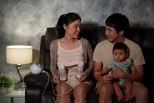 happy father and mother using breast pump machine to pumping milk for infant baby on a sofa in the living room at night