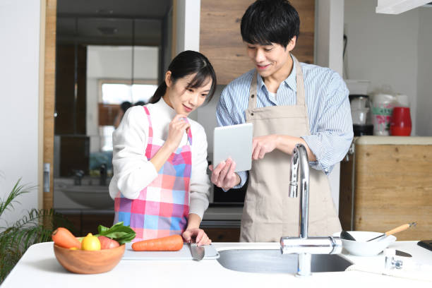 An Asian couple cooks in their home kitchen while checking recipes on a tablet device.