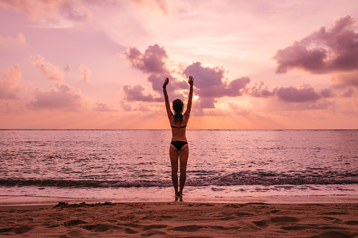 Woman's silhouette with raised arms against calm sunset beach. Freedom and feel good concept