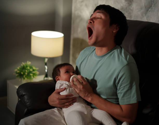 père endormi bâillant et donnant le biberon de lait au bébé sur le canapé dans le salon la nuit - baby yawning asian ethnicity newborn photos et images de collection