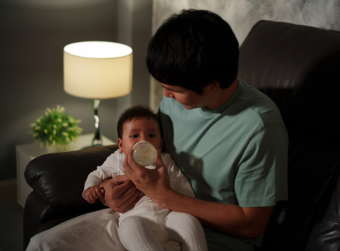 father feeding milk bottle to infant baby on a sofa in the living room at night