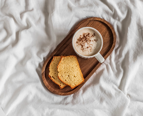 Slice of pound cake, a cup of cappuccino on a wooden tray on the white sheets of the bed. Breakfast in bed