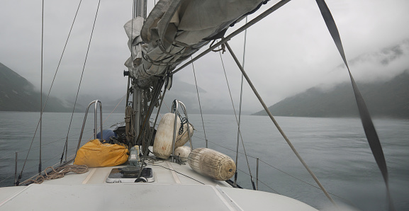Reaching the goal concept, Approaching the misty coast on sail yacht in low visibility on overcast rainy day, fog on the mountains, sail laid on boom, entering Russkaya bay, Kamchatka, panoramic view