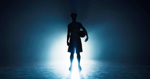Silhouette of male basketball player standing with basketball while playing on sports court during practice.