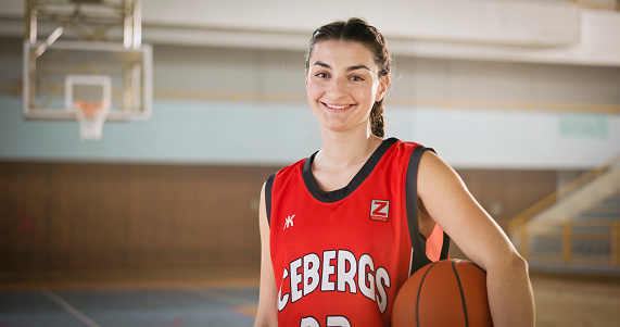 Portrait of smiling young woman standing on sports court with basketball during practice.