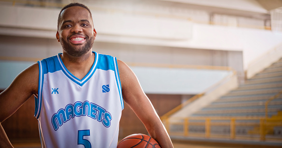 Portrait of smiling male basketball player holding basketball while standing on sports court during practise.