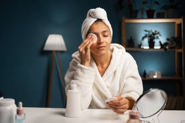 Face care routine in home interior. Hispanic woman in towel looking at mirror and using micellar water with white cosmetic cotton pad for makeup removal Face care routine in home interior. Hispanic woman in towel looking at mirror and using micellar water with white cosmetic cotton pad for makeup removal. Copy space asian night skin care stock pictures, royalty-free photos & images