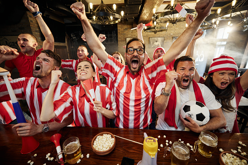 Large group of cheerful soccer fans celebrating the goal of their favorite sports team while watching a game on TV in a bar.